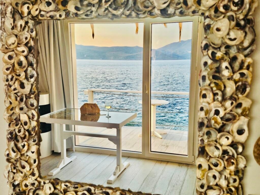 Reflection of a peaceful seaside view through a shell-framed mirror, with a table, wine glass, and serene waters beyond the glass doors
